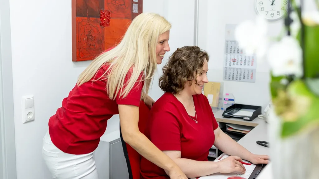 Zwei medizinische Fachangestellte in roten T-Shirts arbeiten lächelnd gemeinsam an einem Computer am Empfangstresen einer Praxis.