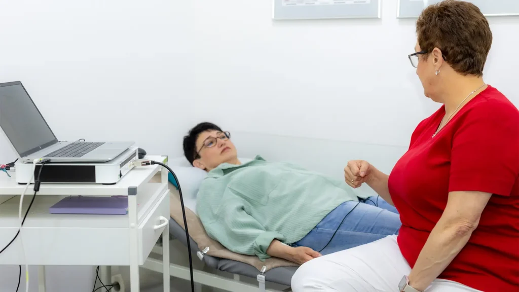 Eine Patientin liegt entspannt auf einer Untersuchungsliege, während eine medizinische Fachkraft in rotem Shirt eine Diagnostik mit angeschlossenen Elektroden durchführt; daneben steht ein Laptop zur Auswertung der Messdaten.