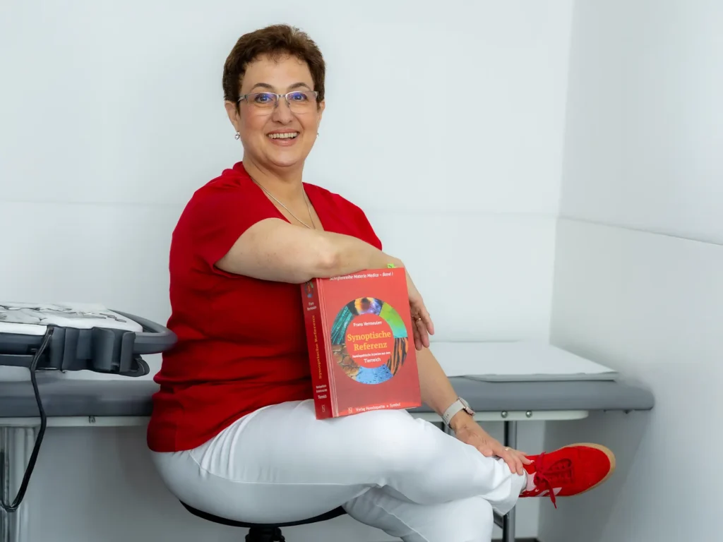 Eine lächelnde medizinische Fachkraft in rotem Shirt und weißer Hose sitzt entspannt auf einem Hocker und hält ein großes medizinisches Fachbuch mit dem Titel „Synoptische Referenz“ auf dem Schoß.