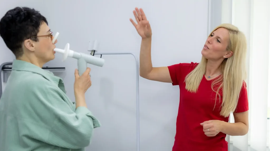 Eine Patientin führt unter Anleitung einer medizinischen Fachangestellten einen Lungenfunktionstest mit einem Spirometer durch.
